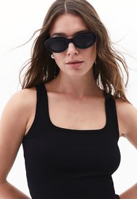 Lunettes de soleil noires en forme ovale avec une finition mate, portées avec un débardeur noir à rayures. Texture lisse, design minimaliste et sans motifs visibles.