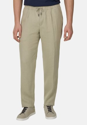 Mężczyzna ubrany w beżowe spodnie z sznurkiem i beżowe tenisówki z czarnymi sznurowadłami, stojący ze swobodnie opuszczonymi rękami na białym tle.