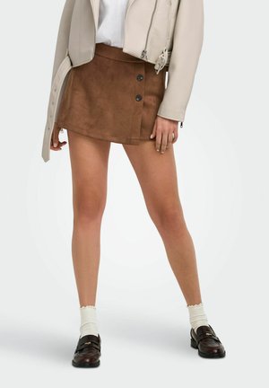 Femme vêtue d'une mini-jupe marron boutonnée, d'une veste beige, d'un haut blanc, de chaussettes blanches et de mocassins marron foncé, debout sur un fond clair uni.