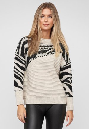 Maglione lavorato a maglia color crema con motivi a zebra neri sulle maniche. Presenta un collo rotondo e polsini a costine. Indossato con leggings neri.