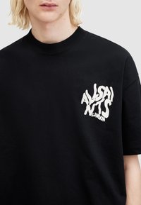 Tricou din bumbac negru cu un logo alb distorsionat pe partea stângă a pieptului. De tip crew neck clasic și cu mâneci scurte. Textură netedă.