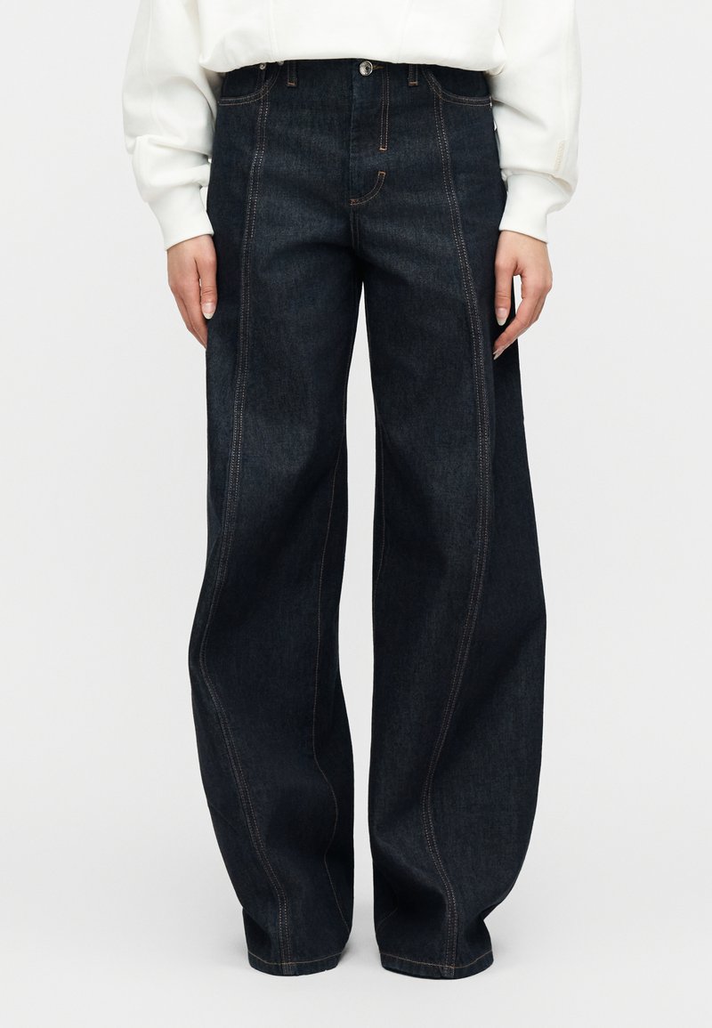 Σκούρα τζιν παντελόνια με φαρδύ πόδι και πορτοκαλί ραφές, φορεμένα με λευκό μπλουζάκι μακριάς μανίκας, τα χέρια χαλαρά δίπλα στο σώμα.