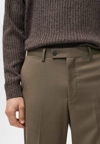 Brązowy prążkowany sweter noszony na oliwkowozielonych, dopasowanych spodniach z płaskim przodem, szlufkami na pasek i pojedynczym zapięciem na guzik.