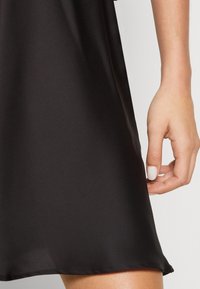 Svart klänning med en slät, glänsande textur och en lätt lyster. Sömmen är rak och en hand håller försiktigt i tyget.