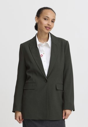 Femme aux cheveux foncés coiffés en chignon, portant un blazer vert foncé sur une chemise blanche boutonnée avec un petit motif rouge.
