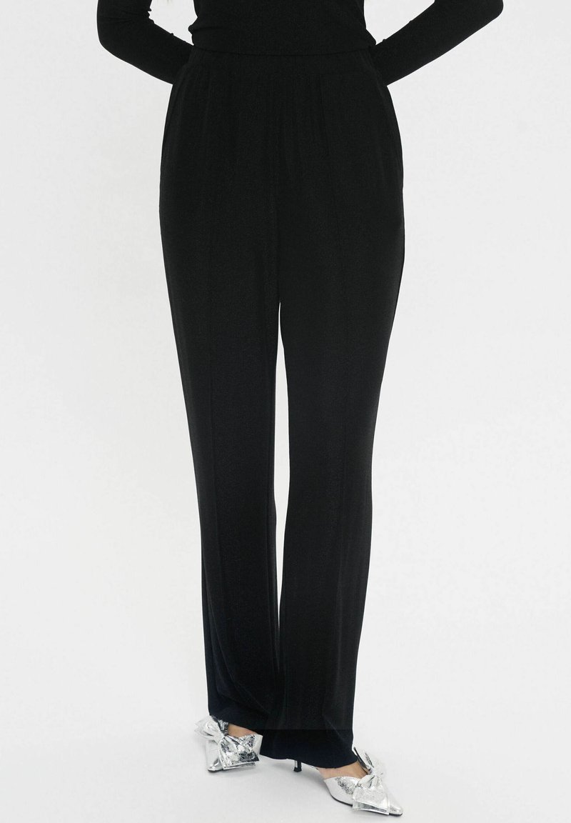 Schwarze, hoch taillierte Hose mit geradem Bein und weichem Stoff. Mit einem elastischen Bund ausgestattet. Kombiniert mit silbernen Absatzschuhen.