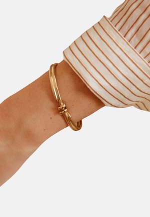 Armbånd i guldfarvet metal med et snoet design og en glat, skinnende overflade, båret på et håndled iført en lysegråstripet skjorte.