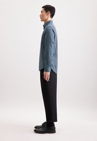 Camicia button-up blu con motivo geometrico, abbinata a pantaloni neri e scarpe formali. Linee pulite, vestibilità sartoriale e silhouette strutturata.