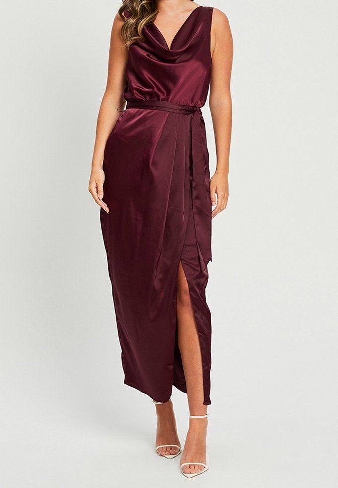 Femme portant une robe portefeuille sans manches en satin bordeaux profond avec col bénitier, fente latérale et ceinture assortie, associée à des talons blancs à bride cheville.