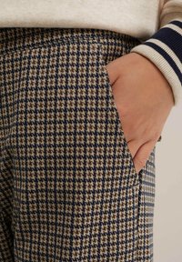 Bordeaux, laivastonsininen ja beige houndstooth-kuvioinen housut, joissa on sivutasku. Kangas on teksturoitu, ja vyötärö on koristeltu raidallisella yksityiskohdalla.