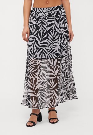 Vrouw draagt een zwart-witte doorzichtige maxi rok met bladmotief, met een onderrok eronder en zwarte sandalen met blokhakken.