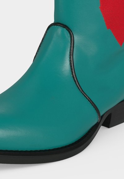 Δερμάτινο μποτάκι τυρκουάζ με μαύρη σόλα και λεπτομέρεια ραφής, με μικρό κόκκινο ύφασμα κοντά στην κορυφή και χαμηλό τακούνι.