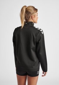 Zwart sportjack met hoge kraag, lange mouwen en witte chevronaccenten op de schouders. Glad textuur, aansluitend ontwerp en logo-detail.
