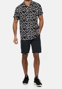 Korte mouwen, knoopsluiting shirt met een zwart-wit geometrisch patroon; gecombineerd met opgerolde donkere shorts en zwarte sneakers.