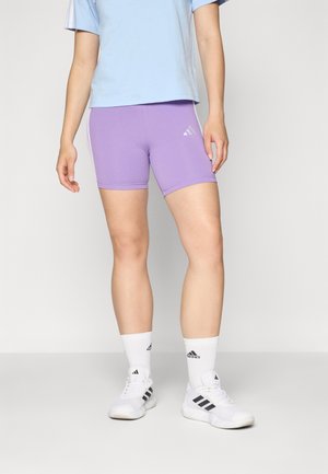Violeta sporta šorti ar baltām sānu svītrām un Adidas logotipu. Valkāti ar gaiši zilu kreklu un baltām kurpēm ar melniem akcentiem.