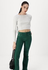 White ribbed crop top with scalloped hem, paired with green flared pants. Model holds a small black handbag. Minimalist design and clean lines.