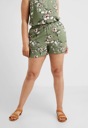 Persona que lleva una blusa verde sin mangas con flores y pantalones cortos a juego con cintura elástica, combinados con sandalias marrones con los dedos al aire sobre un fondo claro.