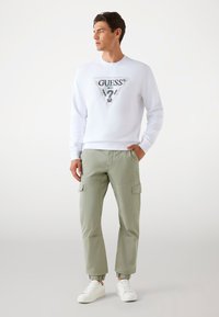 Weißer Baumwoll-Sweatshirt mit einem dreieckigen schwarzen und silbernen "GUESS"-Logo. Grüne Cargo-Hosen mit elastischen Bündchen und Seitentaschen. Weiße Sneakers.