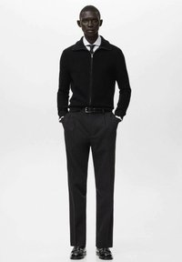 Czarny sweter na zamek z prążkowaną fakturą, zestawiony z dopasowanymi czarnymi spodniami i skórzanymi butami. Widoczna biała koszula i krawat.