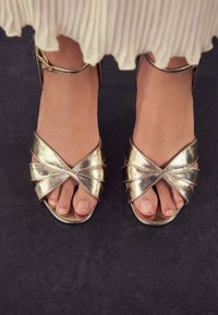 Sandali con tacco dorato metallizzato e cinturini incrociati. Design aperto sulla punta e cinturino alla caviglia visibile. Texture liscia con finitura lucida.