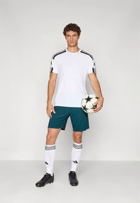 Camiseta atlética branca com riscas pretas, calções azul-petróleo, meias brancas com detalhes pretos e chuteiras pretas, segurando uma bola de futebol preta e branca.