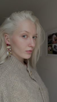 Objemný béžový pletený svetr s vysokým límcem, který ozdobují visací náušnice s červenými a průhlednými korálky, na neutrálním pozadí.