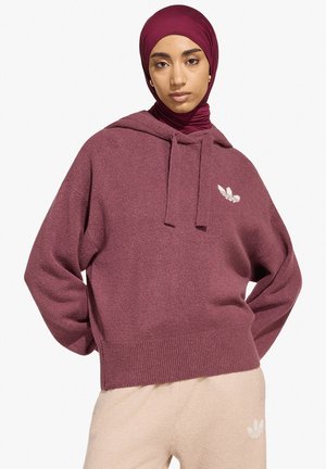 Giovane donna che indossa una felpa con cappuccio color bordeaux, hijab abbinato e pantaloni beige con loghi bianchi, in piedi con le mani sui fianchi su sfondo bianco.