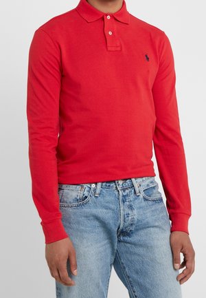 Homme portant un polo rouge à manches longues avec col boutonné et jean bleu clair, debout devant un fond uni.