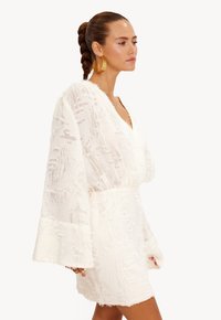 Witte jurk met een textuur van bloemenpatroon, diepe V-halslijn en uitlopende mouwen. Gemaakt van doorschijnend materiaal, met een getailleerd detail in de taille.