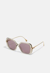 Gafas de sol con montura grande y transparente y lentes morados. Las varillas de metal dorado presentan acentos decorativos cerca de la bisagra.