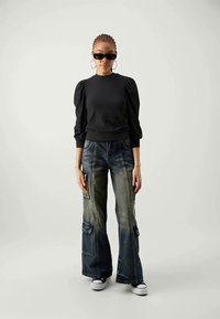 Sieviete ar pītām matiem, ģērbusies melnā puff-sleeve topā, izbalējušās platās kravas biksēs, melnās saulesbrillēs, ar riņķa auskariem un baltajiem sporta apaviem, stāv uz balta fona.
