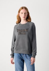Felpa grigia con "TOMMY JEANS" ricamato in una tonalità più scura, colletto rotondo, maniche lunghe e orlo a coste, indossata con jeans blu.