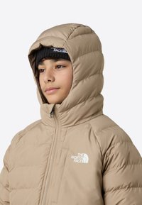 Geacă matlasată bej cu glugă, având textură matlasată, fermoar frontal și logo-ul The North Face alb pe pieptul stâng.