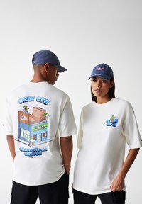 Balta per didelė t-shirt marškiniai su spalvinga grafika ant nugaros, kurioje yra riedlentininkų tema. Mėlyna kepurė, kurią dėvi modeliai, suteikia kontrasto.