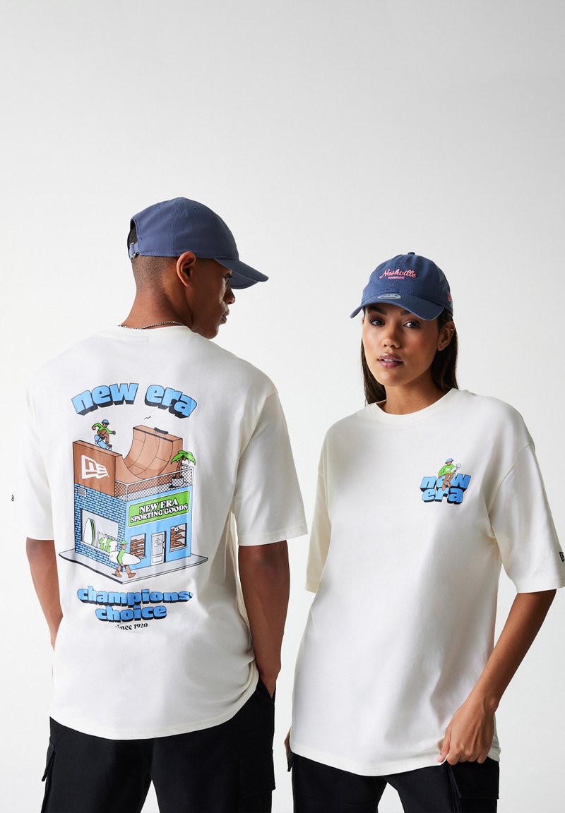 Balta per didelė t-shirt marškiniai su spalvinga grafika ant nugaros, kurioje yra riedlentininkų tema. Mėlyna kepurė, kurią dėvi modeliai, suteikia kontrasto.