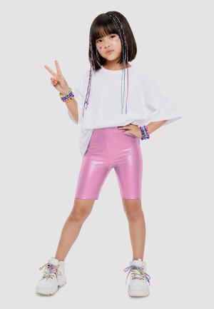 Junges Mädchen trägt ein weißes übergroßes Hemd, glänzende pinke Biker-Shorts, weiße Sneaker mit regenbogenfarbenen Schnürsenkeln, bunte Armbänder und Stern-Gesichtsaufkleber, posiert.