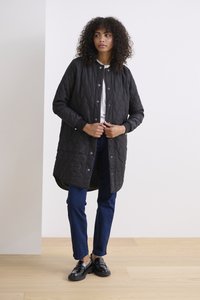 Manteau long matelassé noir avec des boutons-pression, porté sur une chemise blanche et un pantalon bleu, offrant une coupe décontractée et des poches latérales.