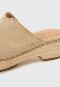 Beige mockaklack med en vävd detalj, en texturerad träsoffa och präglad "TOMS" märkning på sidan. Låg klack med rund design.
