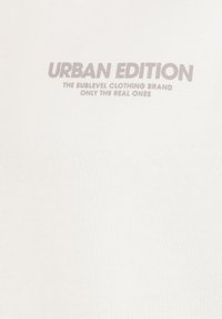 Weißes T-Shirt mit grauem Text, auf dem "URBAN EDITION" steht, sowie zusätzlichen Details darunter, präsentiert auf einem strukturierten Stoffhintergrund.