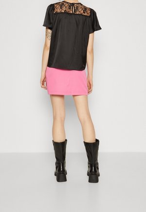 Kobieta ubrana w czarną bluzkę z koronkowym tyłem, różową mini spódniczkę, czarne buty za kostkę, stoi tyłem na jednolitym tle.