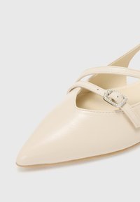 Chaussure en simili cuir beige à bout pointu, avec des brides croisées et une boucle décorative à l'avant. Texture lisse avec un petit talon.