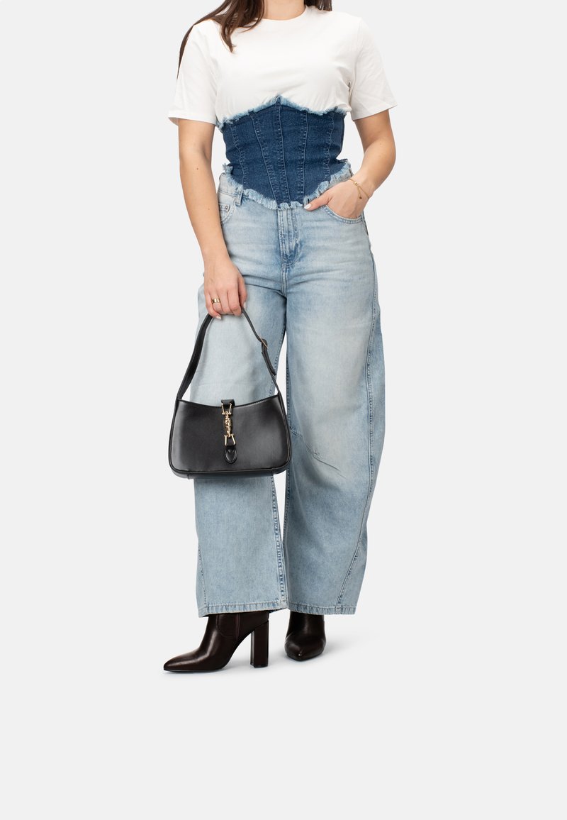 Corsé de mezclilla sobre camiseta blanca, combinado con jeans de pierna ancha de color azul claro y botines negros de tacón. Sosteniendo un bolso negro con herrajes dorados.