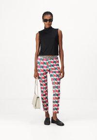 Schwarzes, ärmelloses Oberteil mit hohem Kragen, gemusterte, eng anliegende Hosen in Pink, Grün und Blau mit einem schwarzen Akzentbund, kombiniert mit schwarzen Loafers und Sonnenbrille.