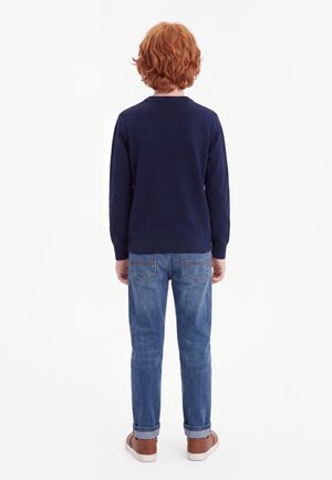 Barn med krøllet rødt hår iført en marineblå sweater, blå jeans og brune sko står med ryggen mod os på en hvid baggrund.