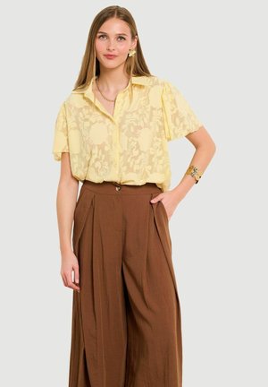 Mladá žena v priesvitnej žltej blúzke s kvetinovým vzorom zastrčenej do hnedých širokých nohavíc s vysokým pásom, so zlatými šperkami a neutrálnym pozadím.