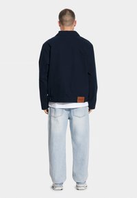 Granatowa bawełniana kurtka z klasycznym kołnierzem, prezentowana na osobie w białym t-shircie i jasnoniebieskich, luźnych dżinsach. Brązowa skórzana metka na plecach.