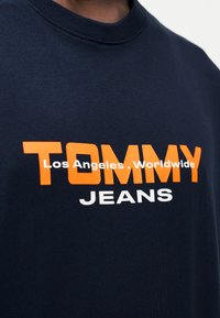 Μπλε βαμβακερό t-shirt με μεγάλο πορτοκαλί κείμενο «TOMMY», μικρότερο λευκό «JEANS» και «Los Angeles, Worldwide» ενδιάμεσα. Με στρογγυλή λαιμόκοψη.