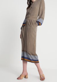 Langes, gemustertes Kleid mit Knopfleiste, braunem Grundton, blauen und orangen Akzenten und tailliertem Schnitt. Kombiniert mit beigen Blockabsatzsandalen.
