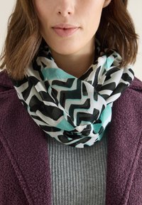 Infinity scarf made of lightweight fabric featuring a geometric pattern in black, white, and teal. Worn over a gray textured sweater.