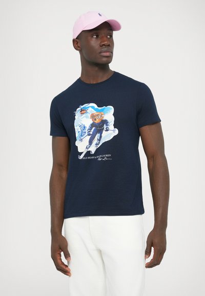 Tamno plava pamučna majica s grafikonom medvjeda na skijanju i helikoptera okruženih snijegom, u kombinaciji sa svijetlo ružičastom kapom.